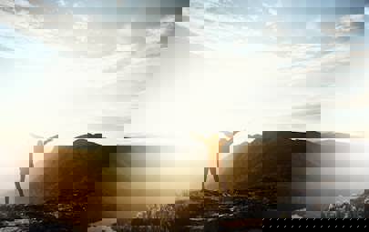 Man standing on a mountain with his arms stretched out while looking at the sun in the distance Man standing on a mountain with his arms stretched out while looking at the sun in the distance