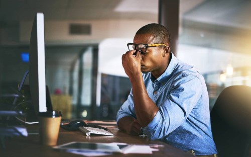 african american male professional sitting at work desk holds nose bridge, in stressed demeanor, worried about staffing challenges