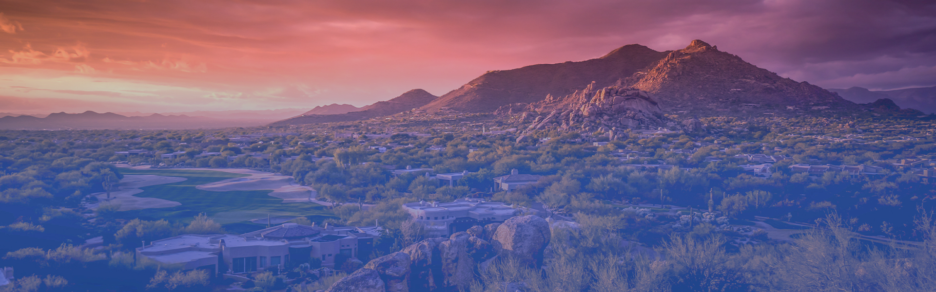 A picturesque sunset over a mountain range, with trees and buildings silhouetted against the orange and pink sky.